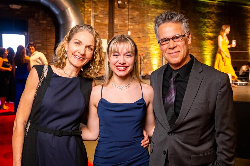 Two women, both wearing navy dresses, smile with a man wearing a black coat and button up shirt.